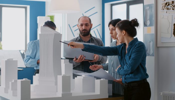 Multi ethnic people working with laptop, tablet and building model for architecture project. Coworkers doing teamwork to design maquette and construction layout with blueprints.
